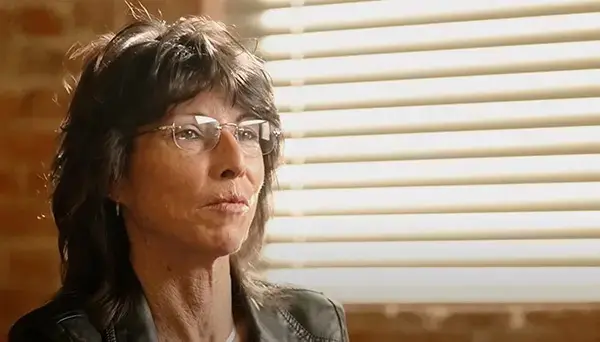 A person with shoulder-length dark hair and glasses sits in a room with natural light streaming through blinds. They wear a dark jacket and appear thoughtful. The background features a brick wall.