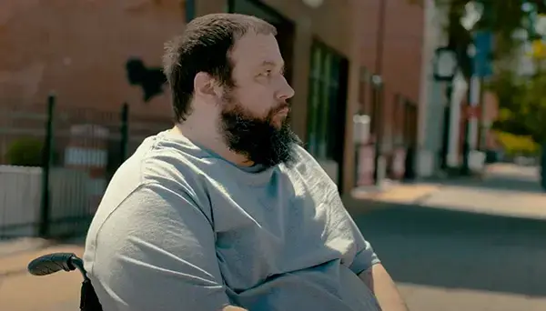 A bearded man in a gray shirt sits in a wheelchair, gazing thoughtfully to the side. The background shows a blurred outdoor street scene with buildings and a few trees.