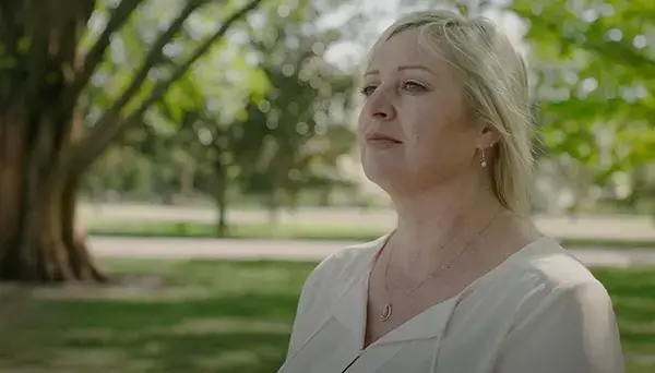 A woman with blonde hair is standing outdoors in a park, wearing a light-colored blouse. She is looking thoughtfully into the distance. The background features green trees and grass, suggesting a sunny day.