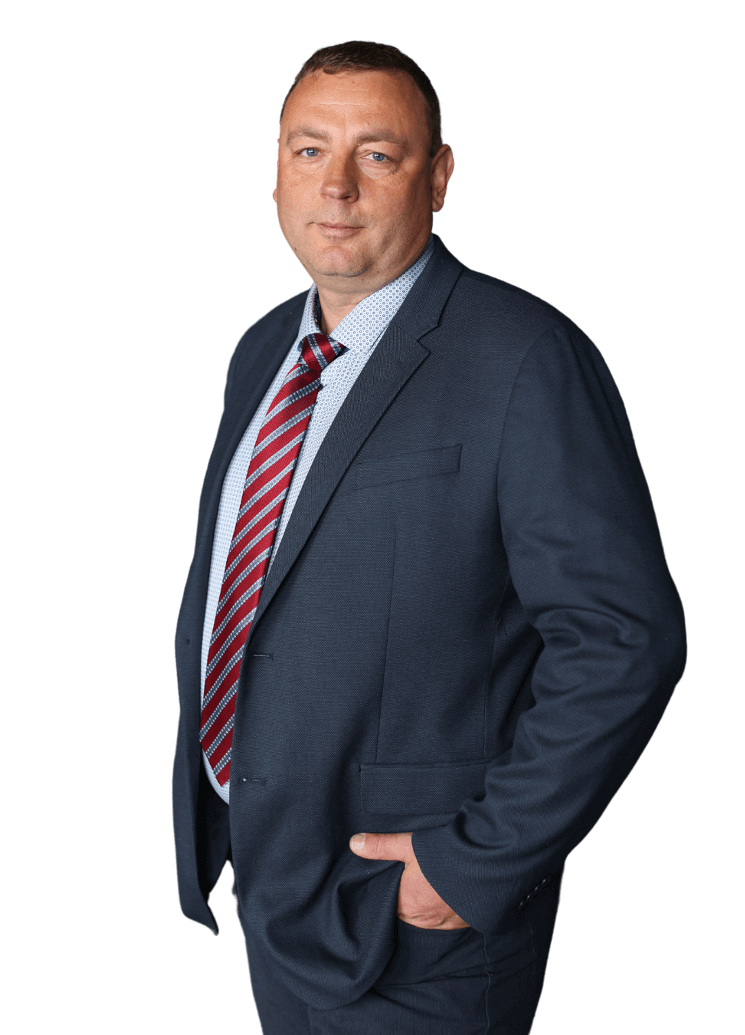 A man in a blue suit with a red striped tie poses confidently, embodying the professionalism of Alex Sutton Immigration & Family Lawyers, with one hand in his pocket against a plain background.