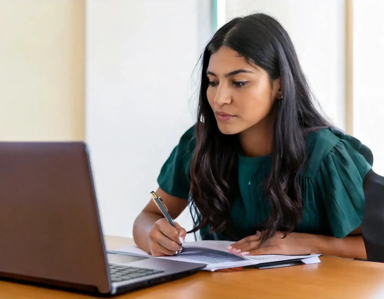 A woman with long dark hair sits at a wooden table, focused on writing in a notebook about the new immigration rules for international students. She is using a laptop, with papers spread in front of her. She wears a green blouse, set against a brightly lit, neutral background.