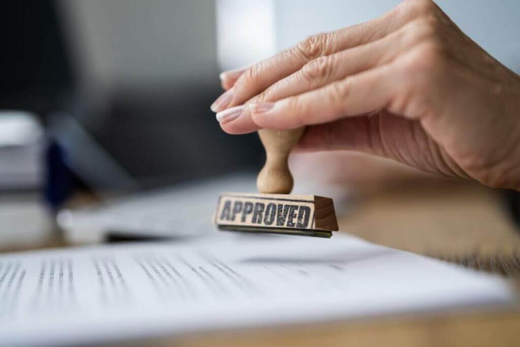 A hand holds a rubber stamp with the word APPROVED in black letters, pressed onto an immigration document on a busy law office desk. In the blurred background, part of a laptop sits, hinting at its essential role for immigration lawyers in Toronto as they manage cases online.