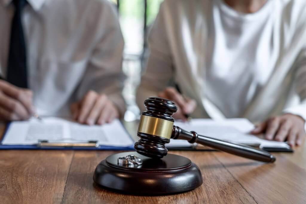 A close-up of a gavel and wedding rings on a table, with two people in the background reviewing documents. Dressed in formal attire, they suggest a legal or court setting related to marriage or divorce proceedings, possibly consulting Ontario divorce lawyers for guidance.