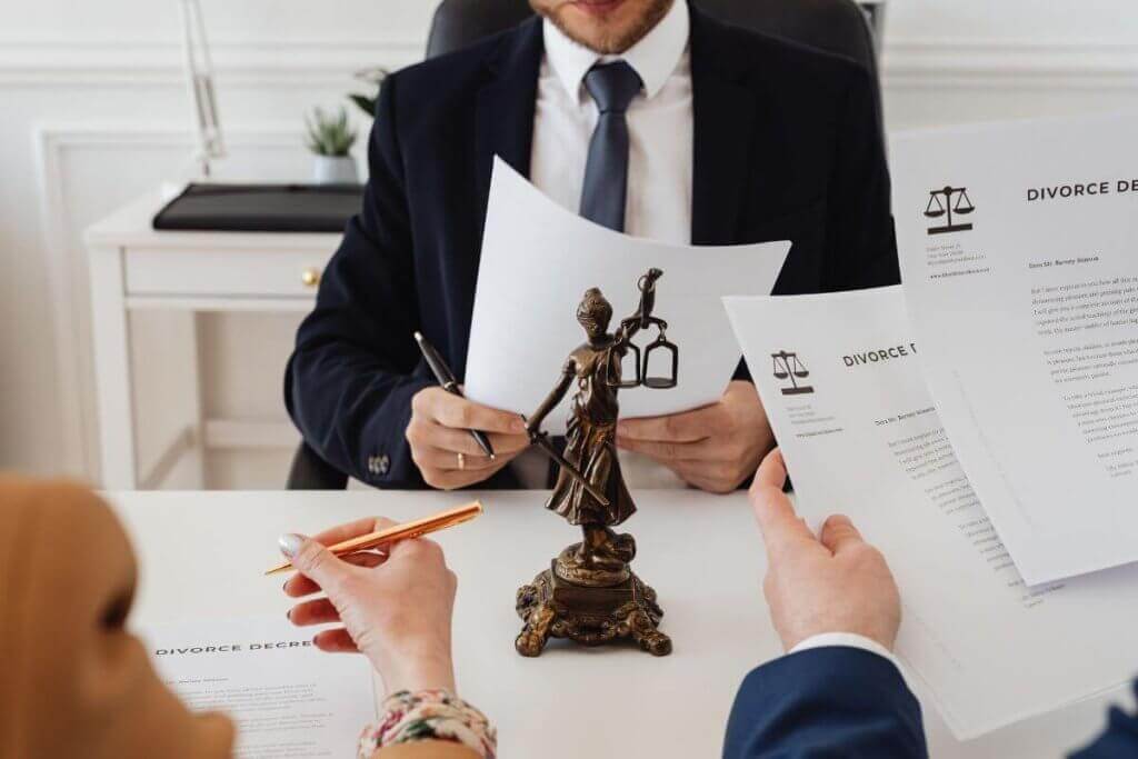 Two people holding divorce papers sit across from a divorce lawyer in a suit, with a Lady Justice statue on the table. Pens and documents are prominent, suggesting a legal discussion or agreement process in Ontario.