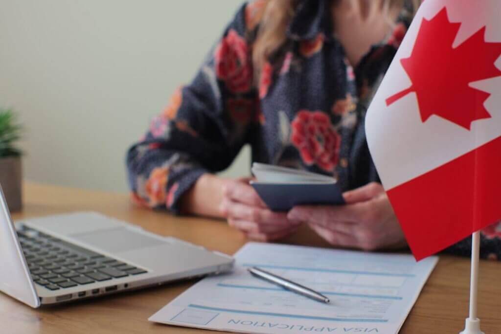 A person holding a passport sits at a table with a laptop and Canadian visa application form, perhaps having consulted immigration lawyers in Toronto. A small Canadian flag is visible in the foreground. The person is wearing a floral shirt, and a small plant is in the background.