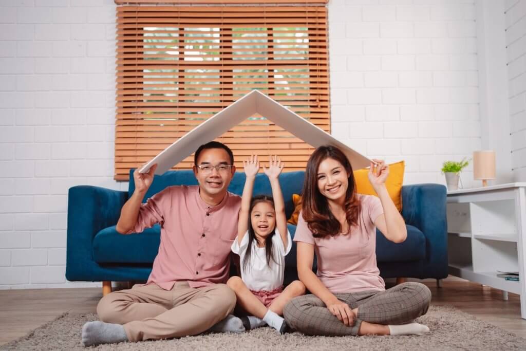 A happy family of three, thoughtfully guided by Family Lawyers Toronto, sits on the floor in a living room. The father, mother, and child hold a large cardboard piece overhead shaped like a house roof. They smile warmly in front of a blue sofa and closed blinds on the window.