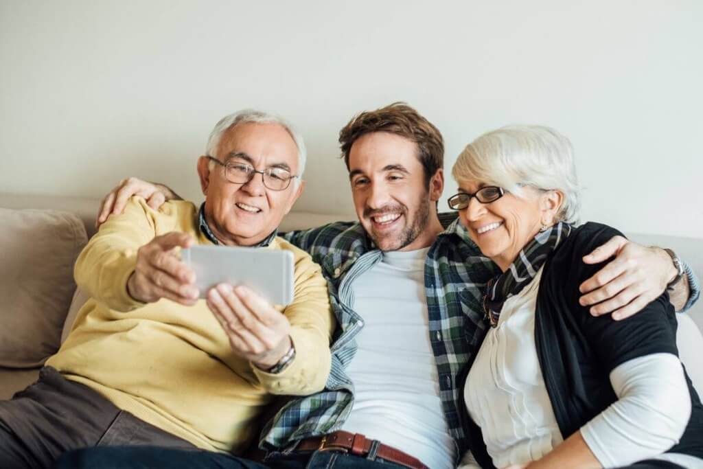 A cheerful older couple and a young man take a selfie on a couch, capturing a happy moment. The older man sports glasses and a yellow sweater while the woman dons specs with a black cardigan. The young man in the plaid shirt smiles brightly, showcasing family vibes reminiscent of joyous meetings at family lawyers in Toronto.