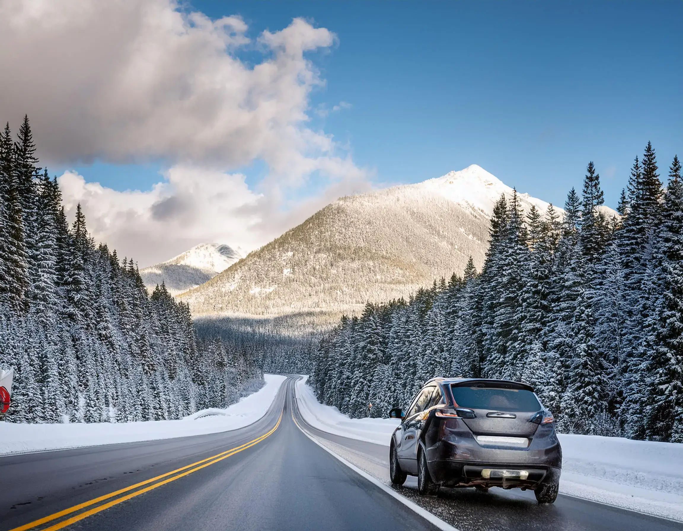 A car travels on a snowy, empty highway flanked by snow-covered trees, leading toward distant mountains beneath a partly cloudy blue sky, much like the paths Ukrainian citizens might explore once visa requirements ease.