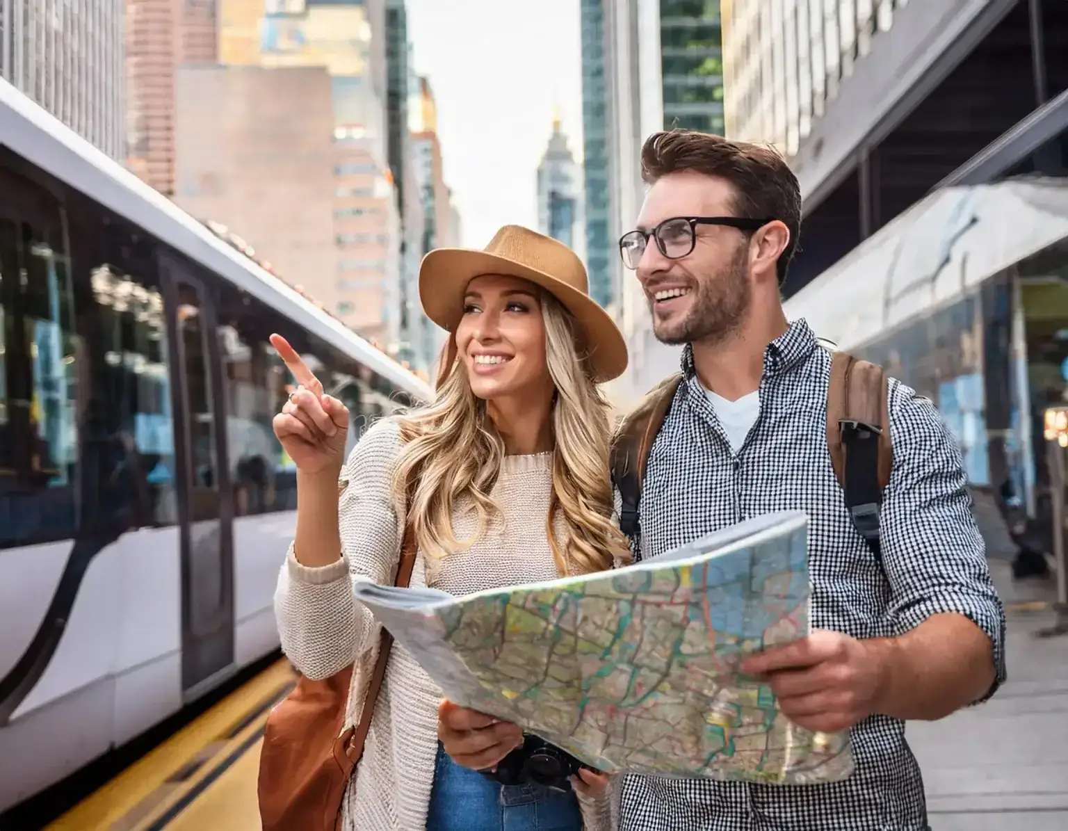 A smiling couple stands at a train station, embodying typical Ukrainian citizens exploring new destinations. The woman in a tan hat and sweater points eagerly at the map, while the man with glasses and a checkered shirt looks on. In the background, a train and tall buildings rise majestically.