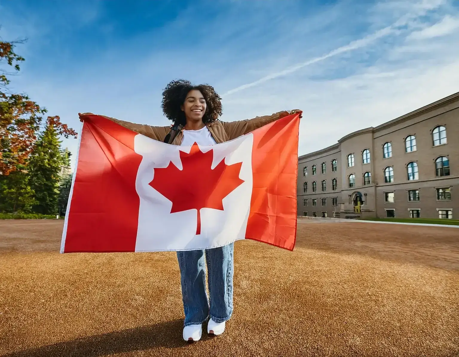 A person celebrating Canada Immigration holds a Canadian flag, standing proudly on a dirt field with trees to the left and a large building to the right under a vibrant blue sky.