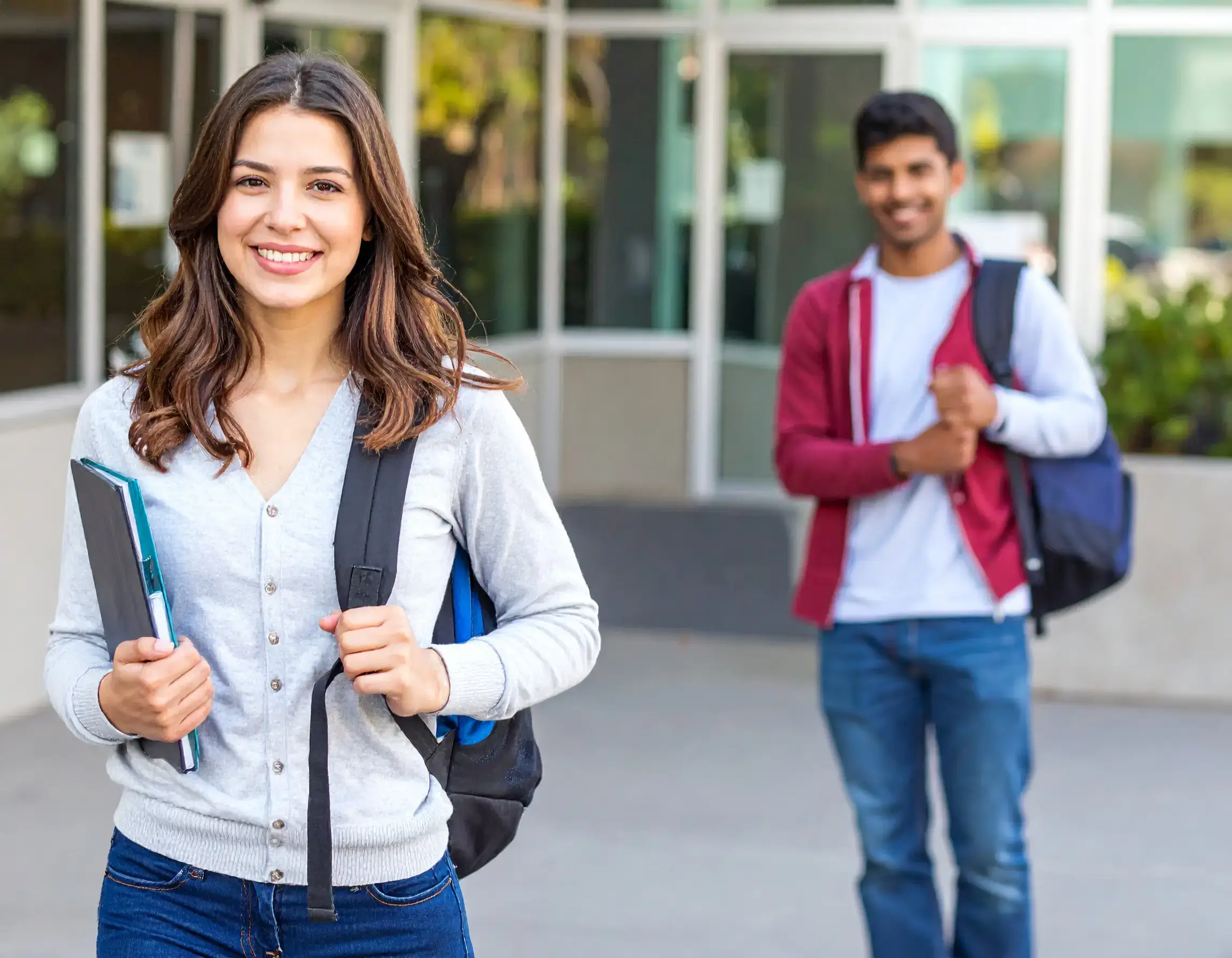 A young woman smiling with books and a backpack stands outside a building, while a young man smiles in the background. Both appear to be students ready to launch their dream career, possibly benefiting from Post Graduate Work Permit Canada policy changes.