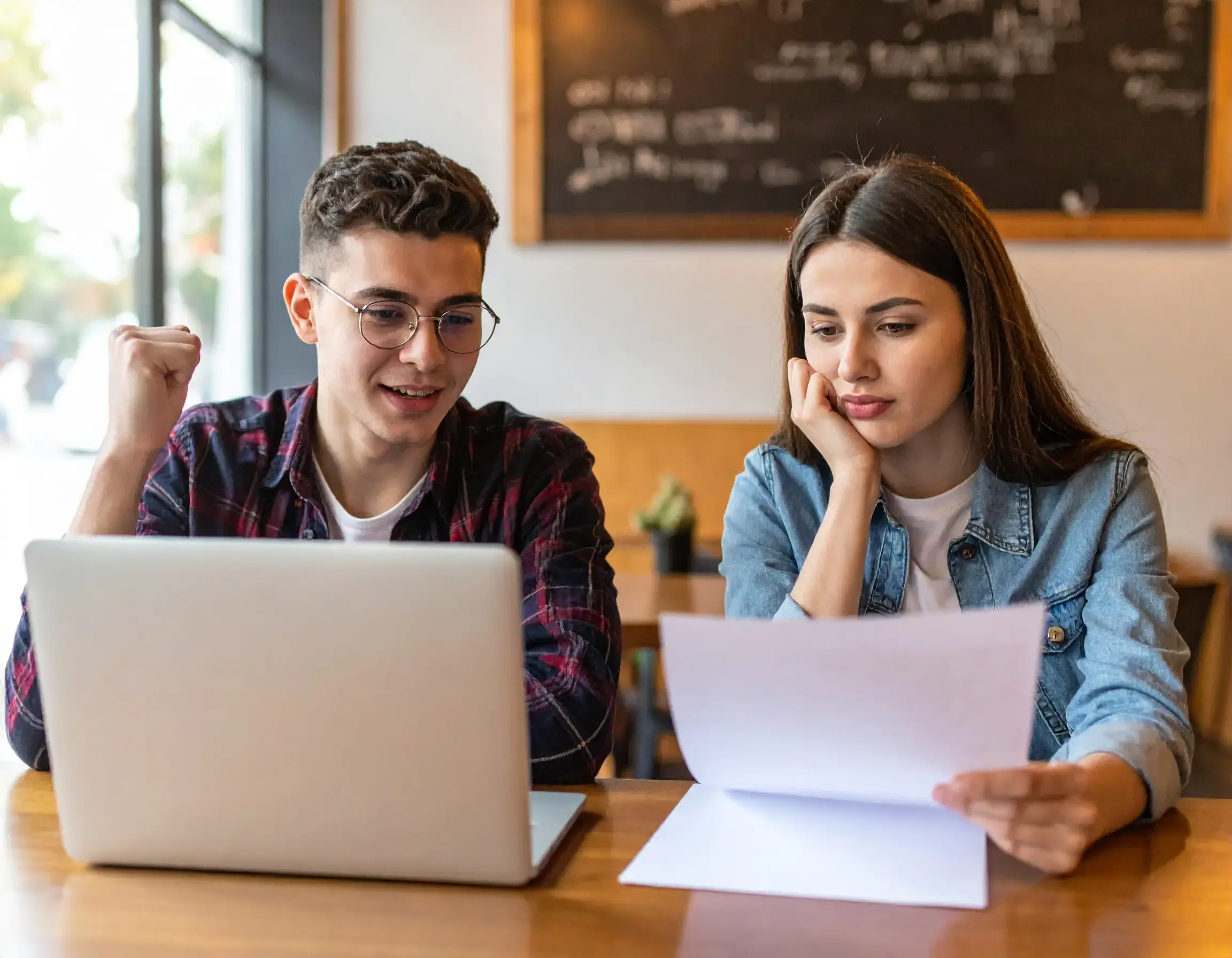 Two young adults sit at a table with a laptop. The person on the left smiles and raises a fist in excitement over their dream career, while the other reviews a document, thoughtful about Post Graduate Work Permit Canada options in a casual indoor setting.