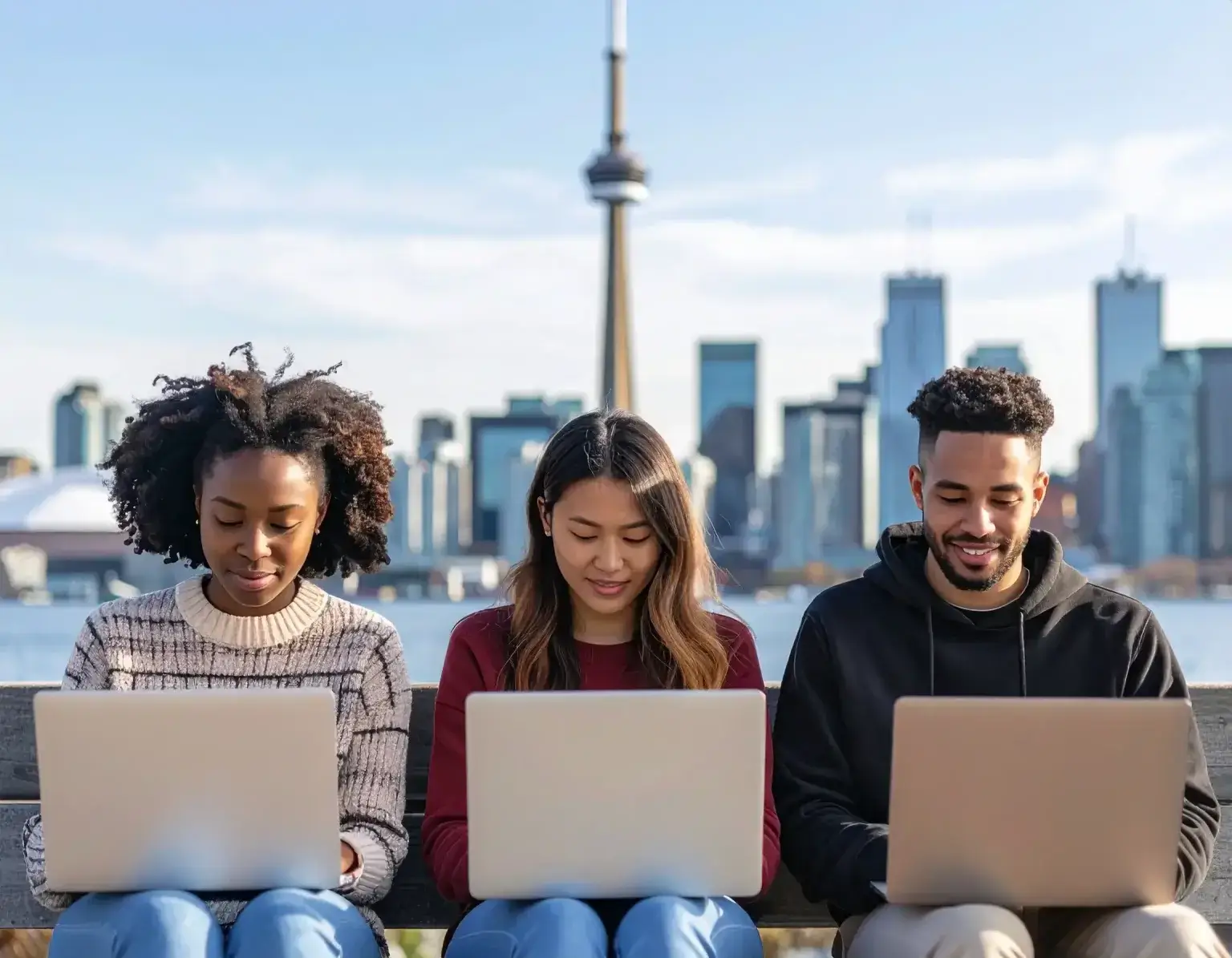 Three young adults sit on a bench outdoors, each working on a laptop, as the city skyline and CN Tower in Toronto rise behind them—a perfect setting to check ITA updates and the latest Express Entry draws under a clear sky.
